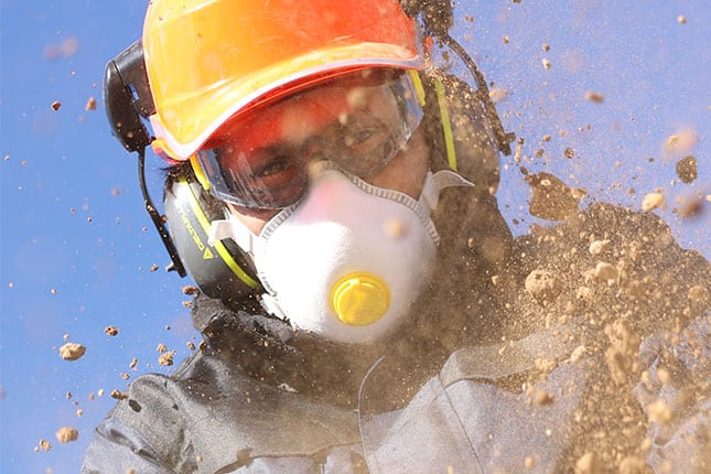 Ouvrier casqué portant un masque anti-poussière FFP à valve et des lunettes de protection, dans un nuage de poussière de chantier